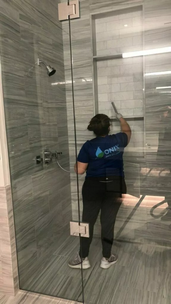 A person wearing a blue "Onix Systems" t-shirt cleans the inside of a glass shower enclosure using a squeegee.