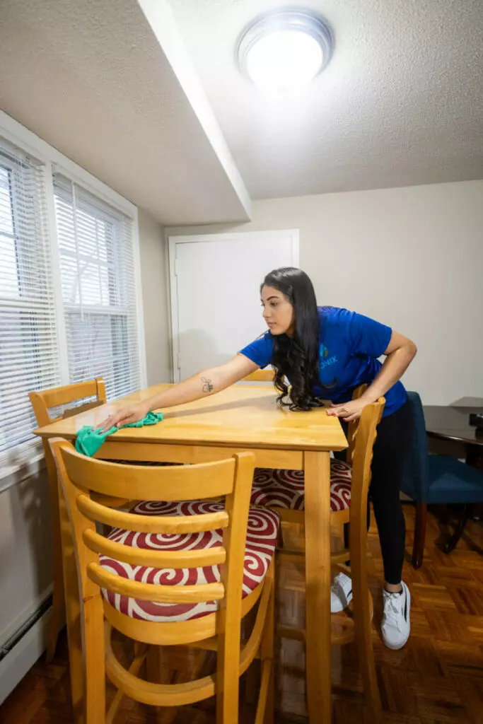 A woman in a blue shirt is wiping a wooden dining table with a green cloth in a small, well-lit room with a wooden floor and a window with blinds.