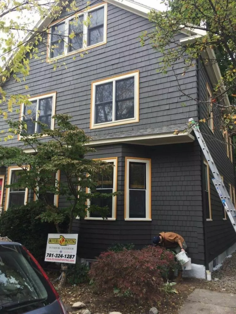 A painter works on the exterior maintenance of a three-story house with gray siding and yellow-trimmed windows. An "Oliveira Painting" sign with a phone number is placed in the front yard, while a ladder leans against the house.