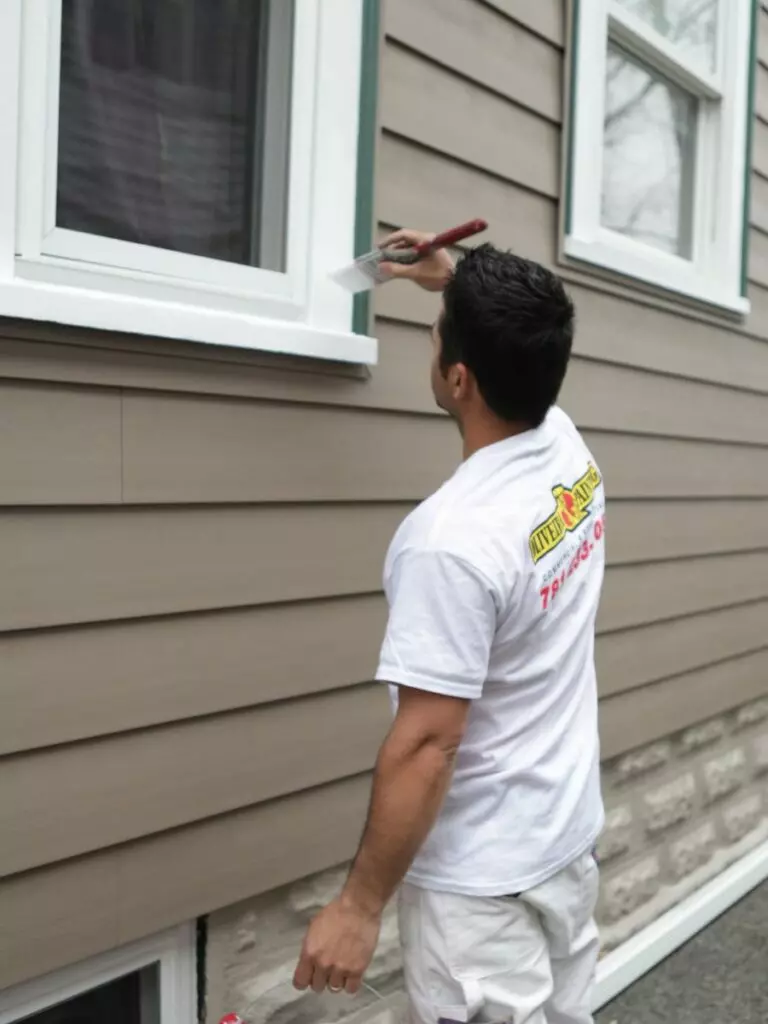 A person in a white shirt and shorts paints a window frame on a beige house with a brush, ensuring meticulous home maintenance.