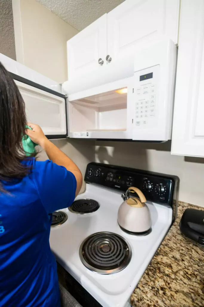 A person in a blue shirt is opening a microwave door above a stove with a kettle on it in a kitchen setting.
