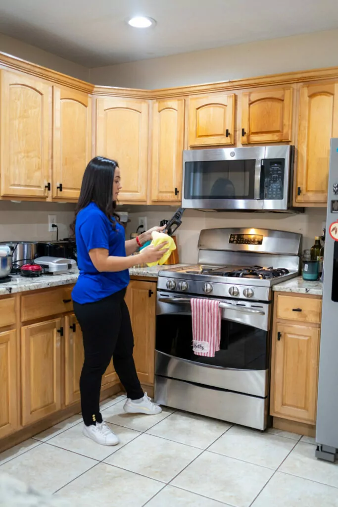 A woman in a blue shirt and black pants is standing in a kitchen with wooden cabinets, holding a pan and a yellow cloth near a stainless steel stove and microwave.
