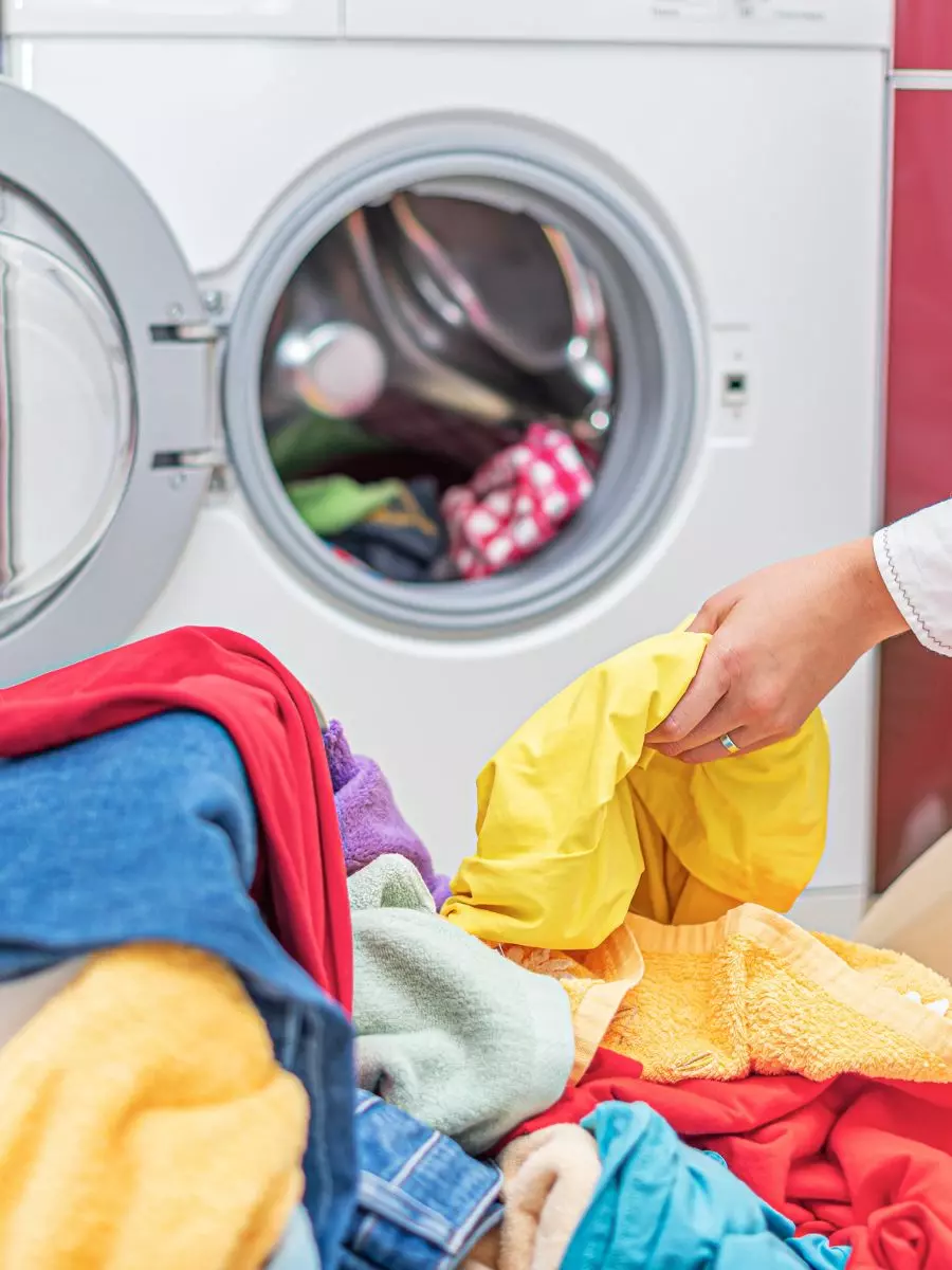 A person is loading colorful clothes into a front-loading washing machine as part of their interior maintenance routine. The machine's door is open, revealing various garments in the drum and scattered on the adjacent surface.