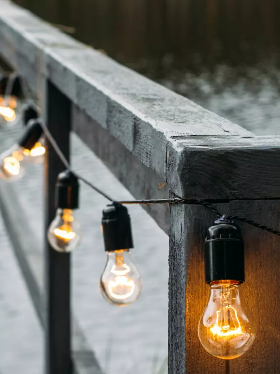 String lights with exposed bulbs are draped along a wooden railing, glowing softly against a dim backdrop, adding a touch of charm without compromising exterior maintenance.
