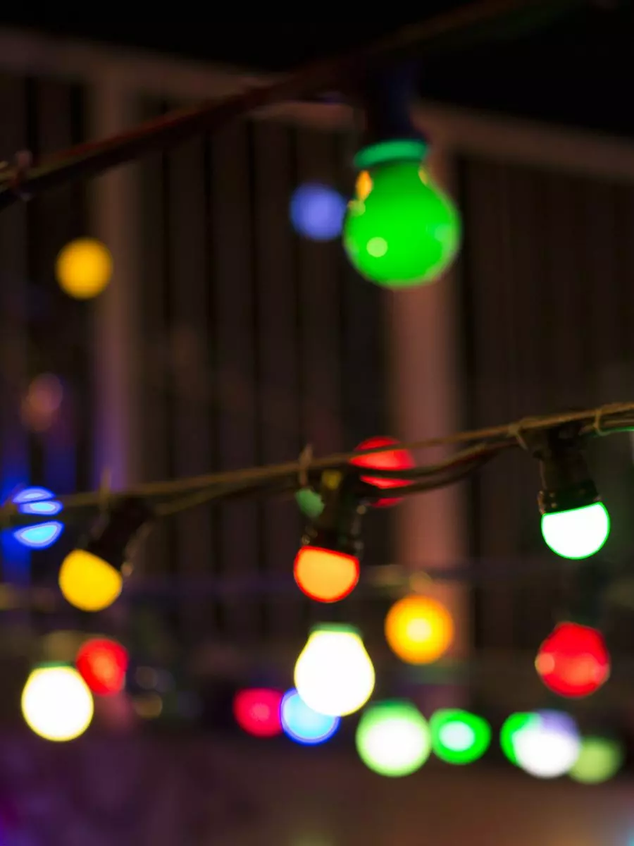 Close-up of a string of multicolored lights hanging outdoors at night, with green, red, yellow, and blue bulbs in focus against a blurred background. Proper exterior maintenance ensures these vibrant lights shine brightly all season long.