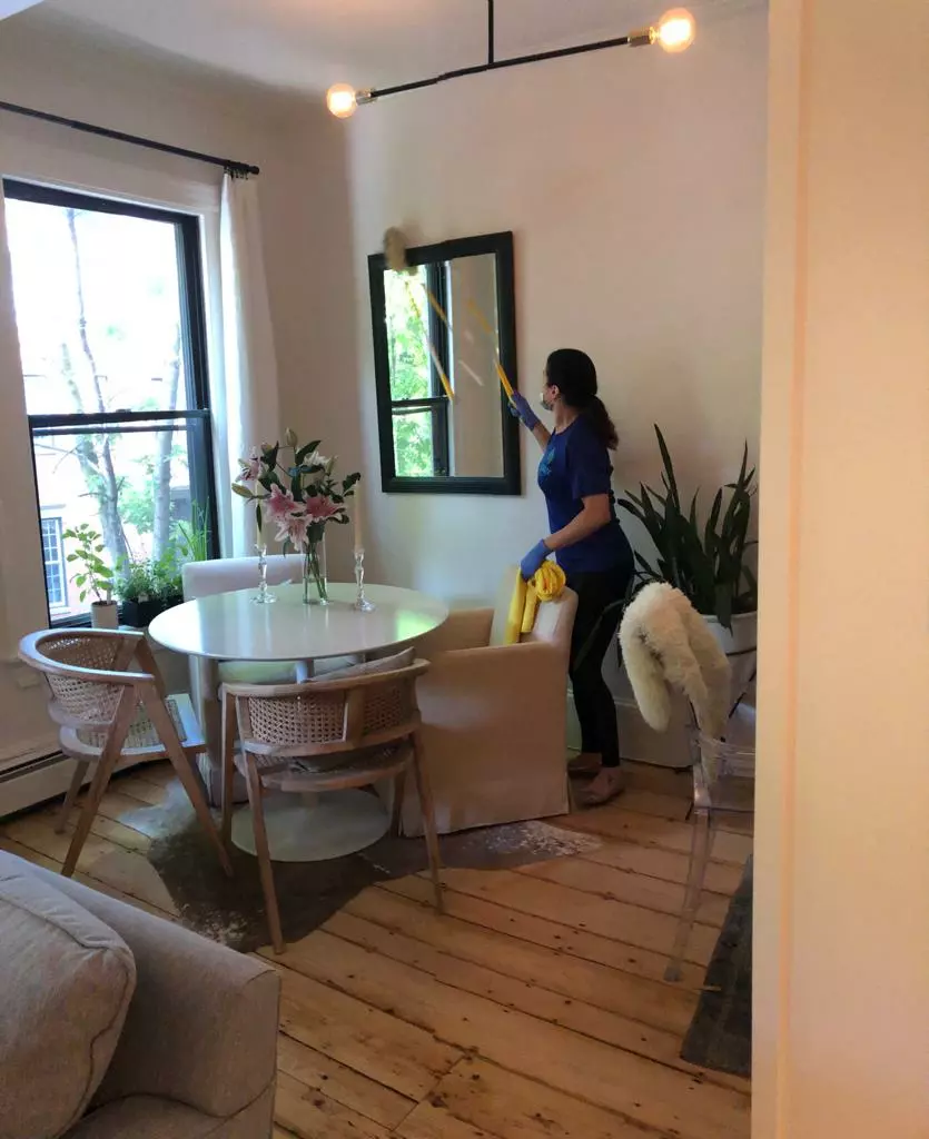 A person in a blue shirt and dark pants is dusting a black-framed mirror with a yellow duster in a dining room with a table, chairs, plants, and a large window.