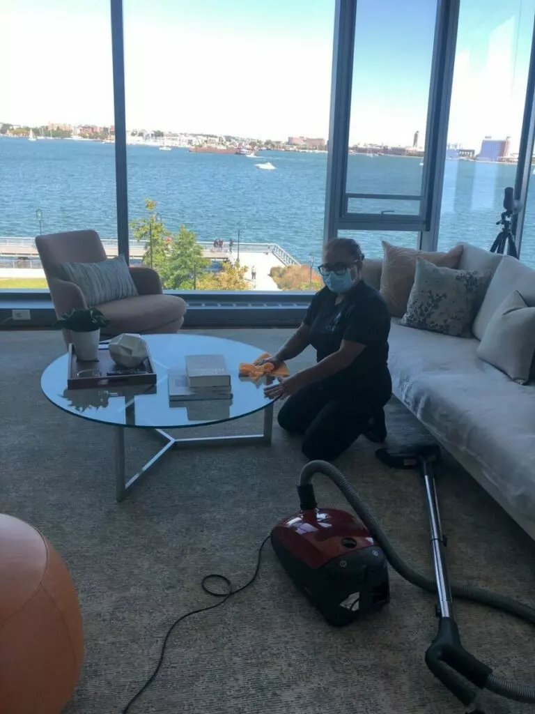Person in a face mask cleaning a glass coffee table in a living room with a vacuum cleaner on the floor. Large windows in the background offer a view of a waterfront scene.