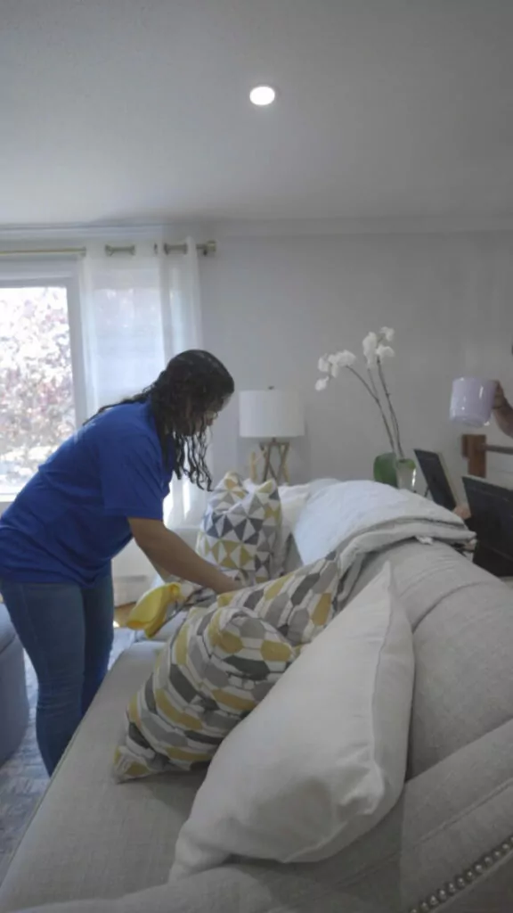 A person in a blue shirt is arranging pillows on a grey couch in a well-lit living room. The room has a window with white curtains, a lamp, and a decorative plant on a side table.