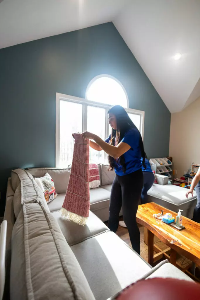 A woman in a blue shirt is folding a red and white blanket on a light gray sofa in a living room with a high, arched window.
