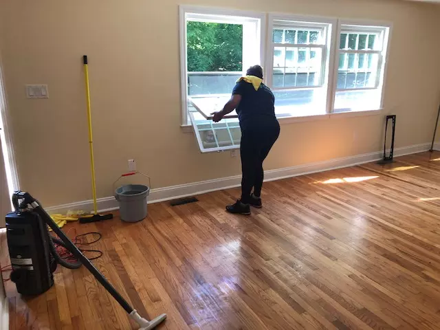 A person is cleaning a large window in a sunlit room with hardwood floors. A vacuum, mop, bucket, and cleaning supplies are visible on the floor.
