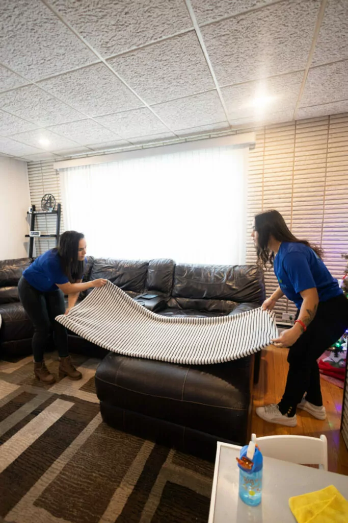 Two people in blue shirts are placing or removing a striped rug on or from a wooden floor in a living room with a dark sofa and a window in the background.