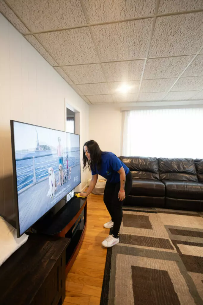 A person in a blue shirt and black pants stands wiping a television screen in a living room with a brown leather couch and a patterned rug.