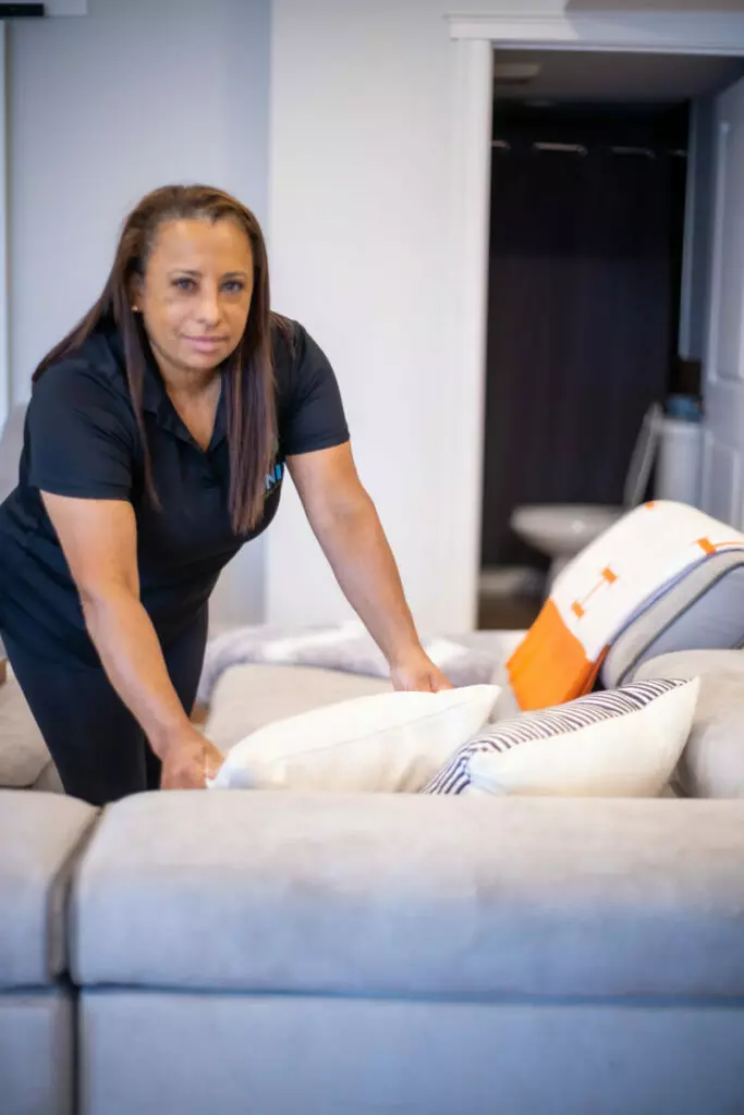 A person adjusts pillows on a grey couch in a brightly lit room.