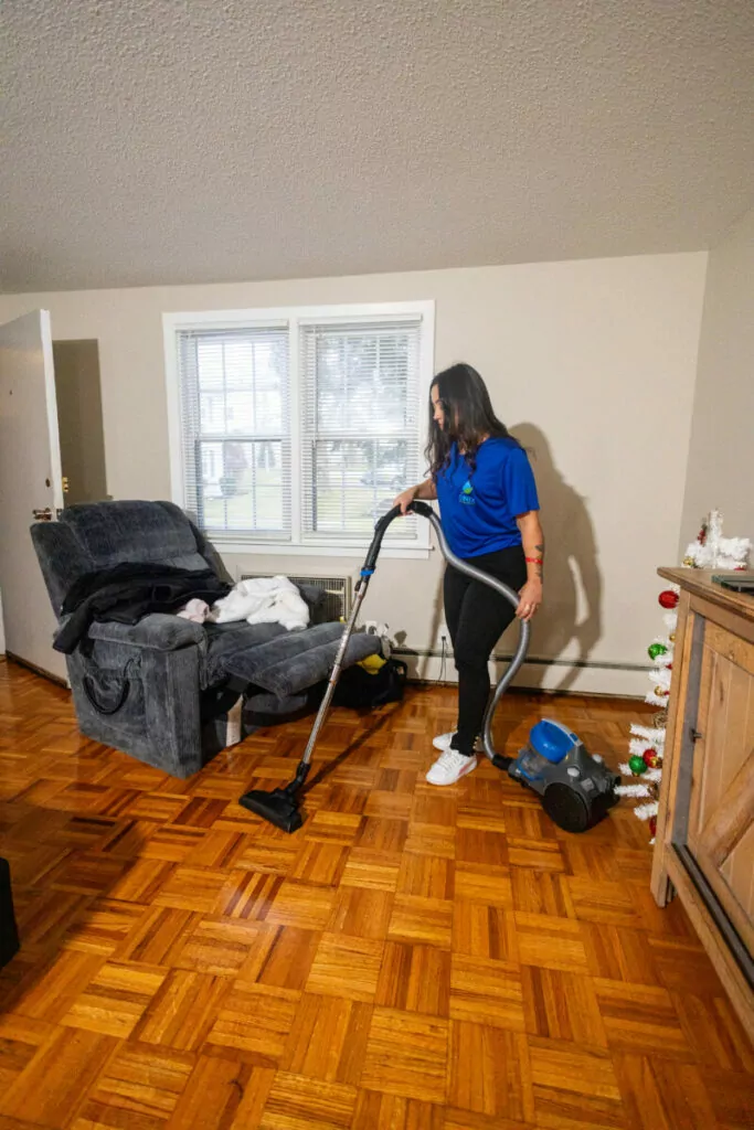 A woman in a blue shirt is vacuuming the wooden floor of a living room with a canister vacuum cleaner. The room has a recliner, a window, and a wooden cabinet with decorations on it.