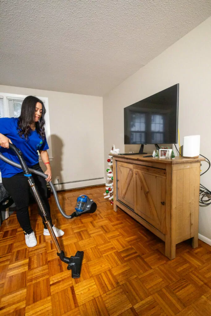 A person in a blue shirt is vacuuming the wooden floor of a living room that has a TV on a wooden cabinet and a small decorated tree next to it.