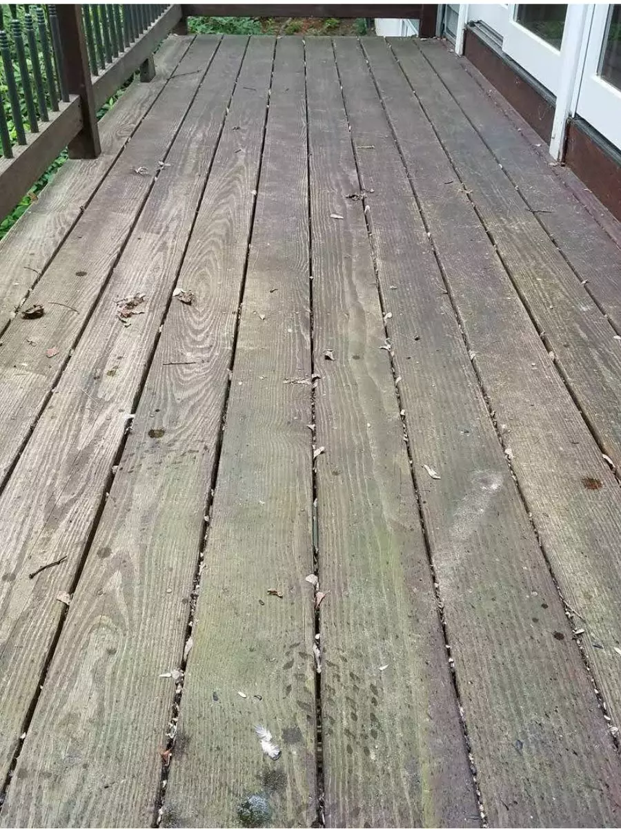 A weathered wooden deck with gaps between the planks, surrounded by a railing, showing signs of dirt and minor debris, is in need of exterior maintenance.