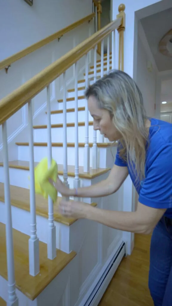 A person in a blue shirt uses a yellow cloth to clean a white staircase railing with wooden steps in a well-lit interior.