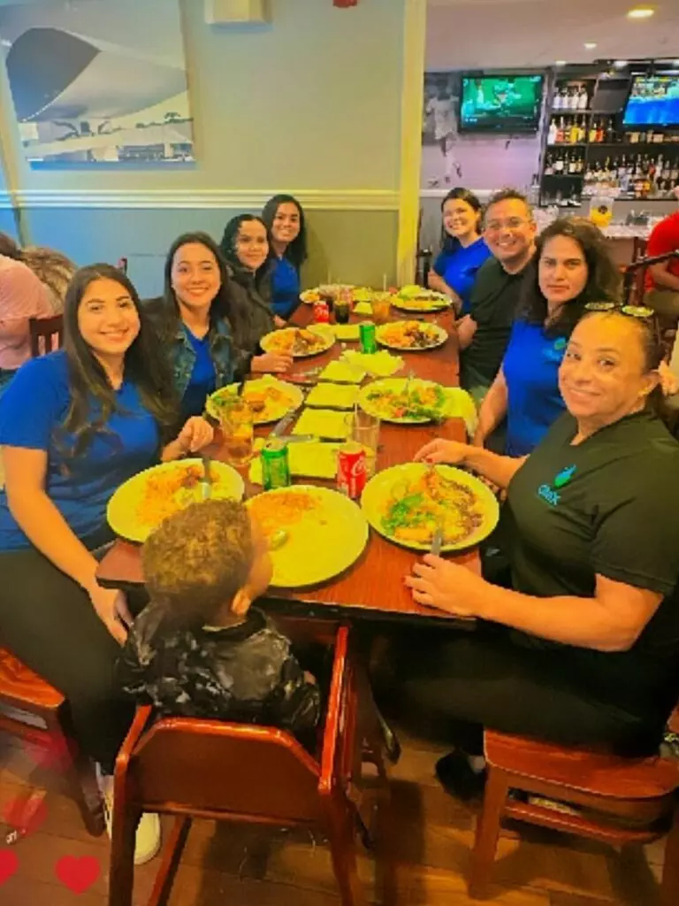 A group of people in blue and black shirts are seated around a table at a restaurant, enjoying a meal together like an artful gallery of companionship. A child is seated in a high chair at the head of the table.