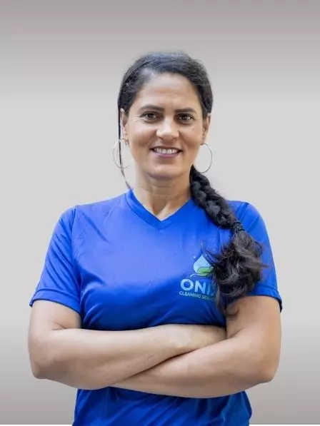 A woman with dark braided hair is smiling and standing with her arms crossed. She is wearing a blue shirt with a logo that says "ONIT Cleaning Services" on it. The background, reminiscent of a gallery wall, is plain and light gray.