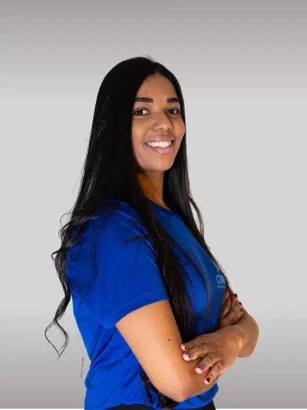A person with long dark hair wearing a blue shirt stands with arms crossed, smiling at the camera against a light grey background, reminiscent of a timeless portrait you might find in an art gallery.
