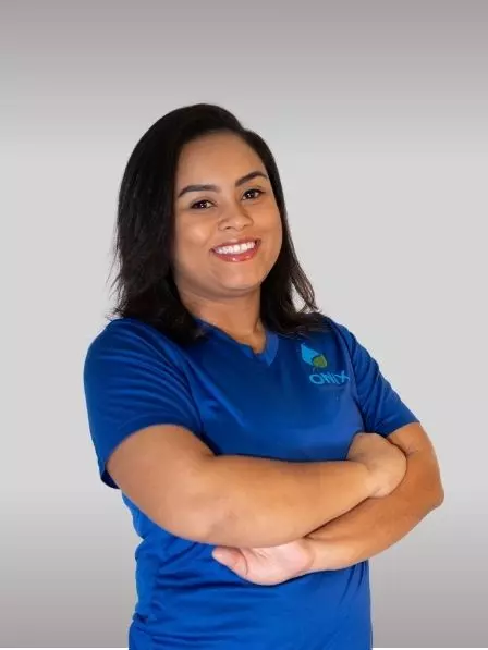A person with shoulder-length hair stands with arms crossed, wearing a blue shirt with a logo on it. The neutral background resembles a minimalist gallery, allowing the focus to remain on their composed demeanor and stylish attire.