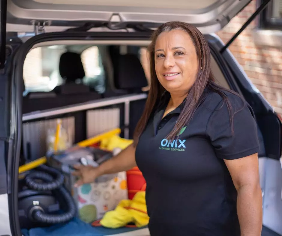 A woman in a black polo shirt labeled "Onix Cleaning Services" stands beside an open car trunk filled with cleaning supplies, resembling a well-organized gallery. She is looking at the camera and smiling.