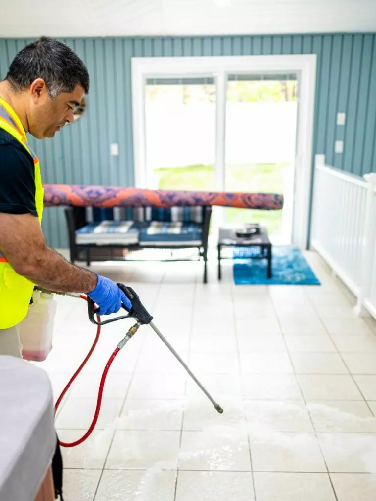 A person in a high-visibility vest is engaged in floor maintenance, cleaning the tiled floor with a hose in a bright indoor space with a blue mat and furniture in the background.
