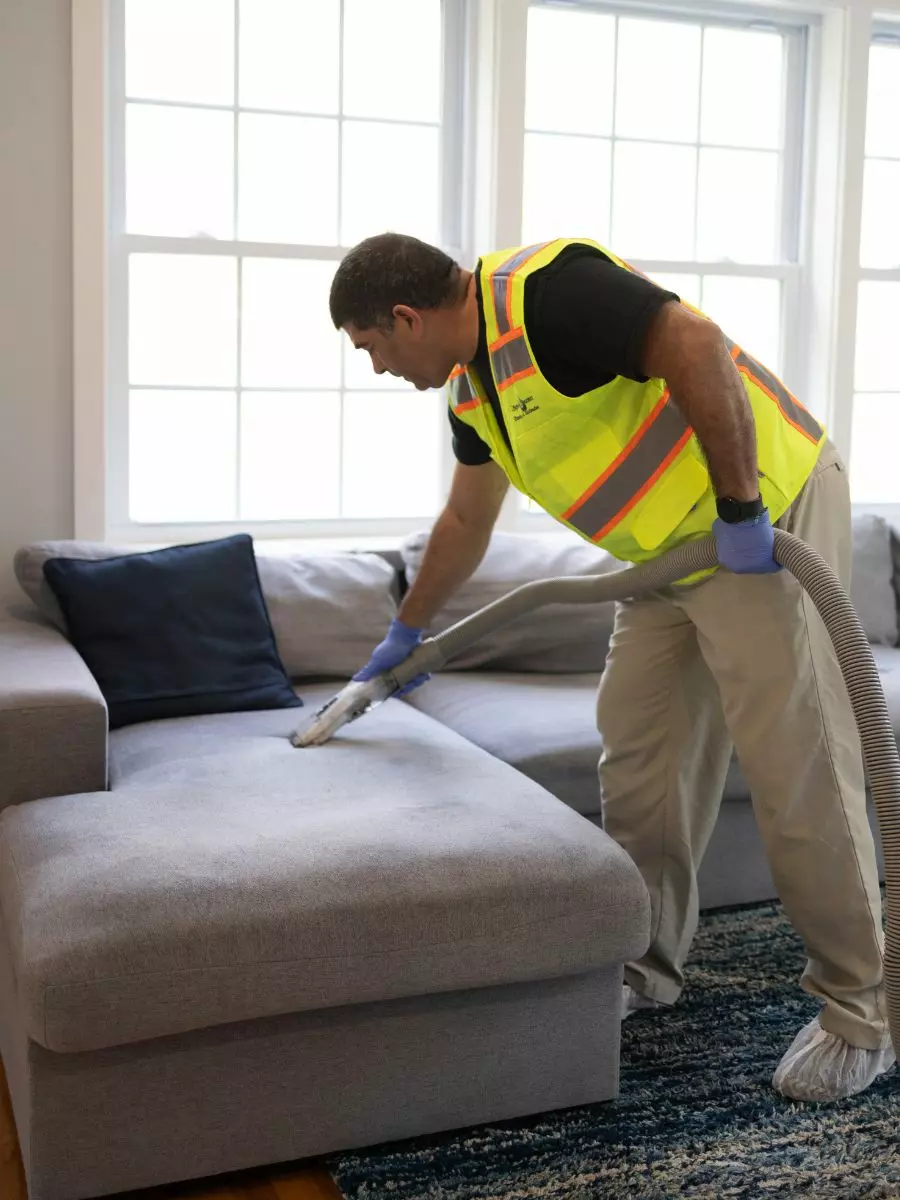 A person in a high-visibility vest and gloves is performing interior maintenance by using a vacuum cleaner to clean a gray sectional sofa in a room with a blue rug and large windows.