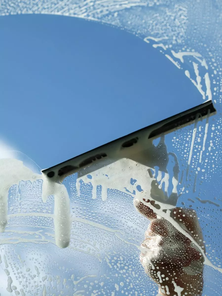 A hand expertly maneuvers a squeegee to clean soapy water from a glass surface, revealing a clear blue sky in this exterior maintenance task.