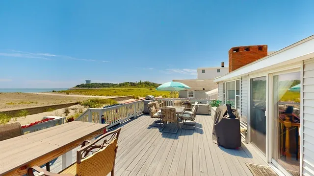A wooden deck with outdoor furniture, a grill, and an umbrella overlooks the beach and ocean under a clear blue sky in charming Winthrop, MA.