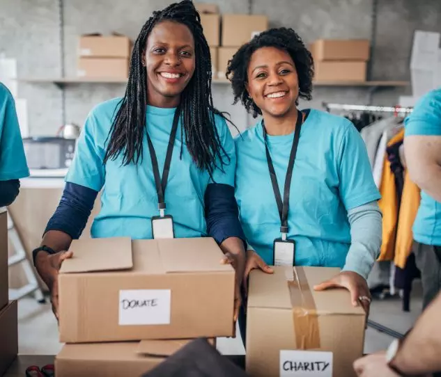 In a room filled with shelves and boxes, two people in blue shirts are smiling as they handle packages labeled "Donate" and "Charity." It's a heartwarming scene of generosity, much like the community spirit found in Winthrop, MA.