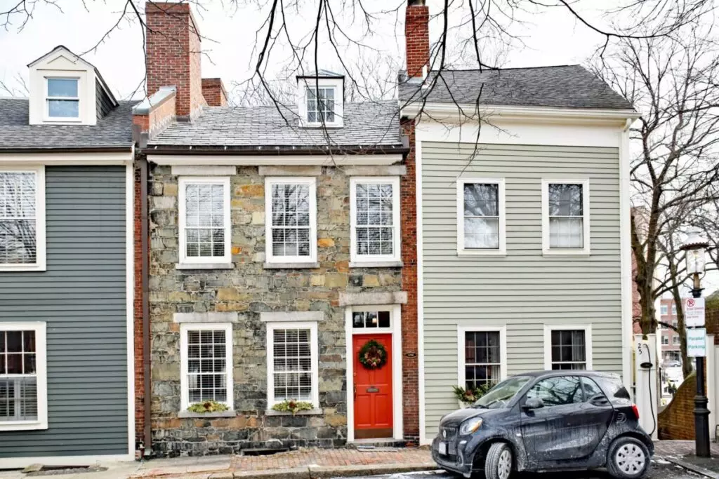 In Winthrop, MA, a row of narrow, old stone and wood townhouses with chimneys is adorned with a red door bearing a wreath. A small car is parked in front on the street, while leafless trees elegantly frame the scene.