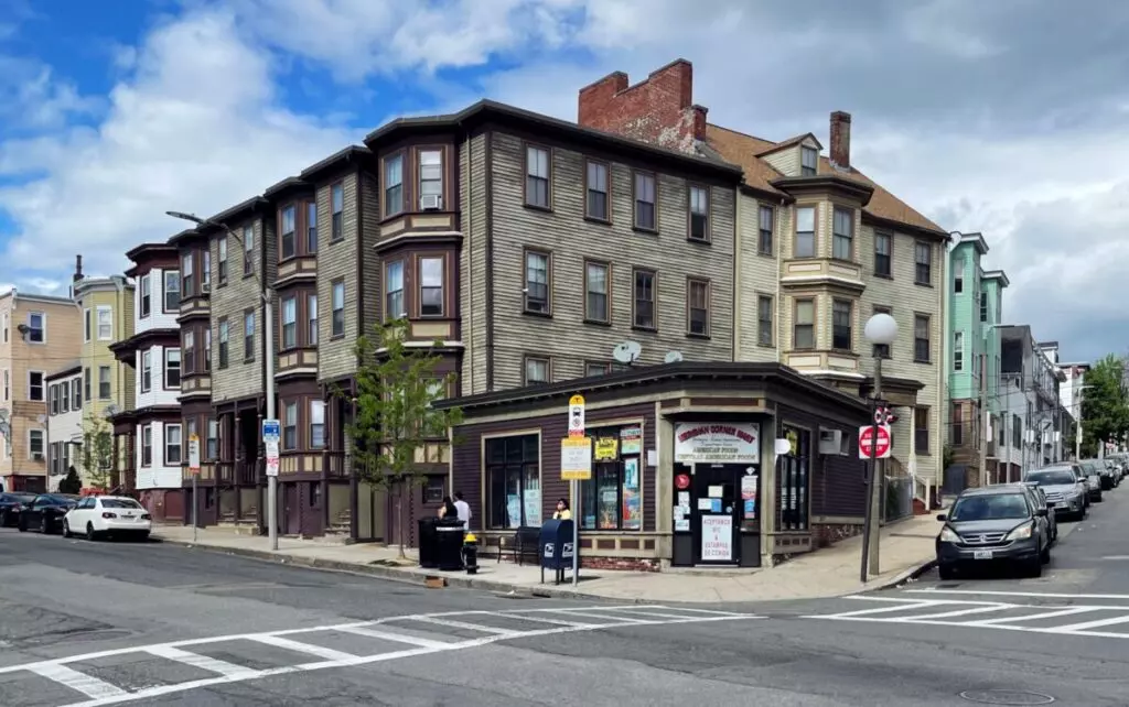 A charming three-story building in Winthrop, MA, features bay windows and a cozy shop on the ground floor at a bustling street corner. The sky is partly cloudy, casting soft shadows over several parked cars lining the streets.
