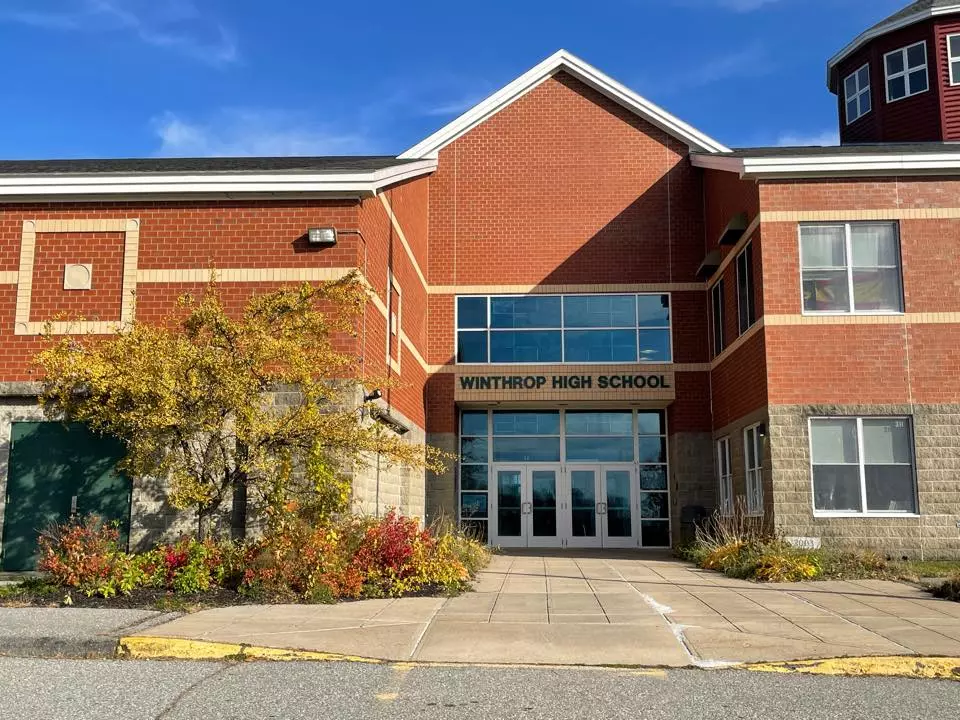The front entrance of Winthrop High School, nestled in red brick, stands proudly under a clear blue MA sky, with vibrant plants adorning the facade.