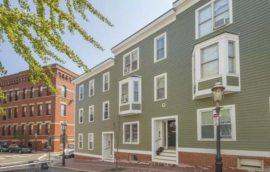 Nestled on a street corner in Winthrop, MA, this green and white townhouse boasts charming bay windows beside a red brick building. A tree to the left and a streetlamp in front complete the picturesque urban scene.