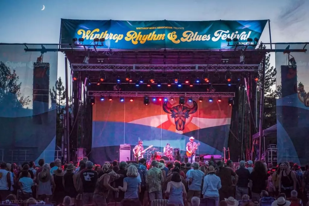 Outdoor concert stage with musicians performing at the Winthrop Rhythm & Blues Festival in MA; the crowd watches under a twilight sky with a crescent moon.