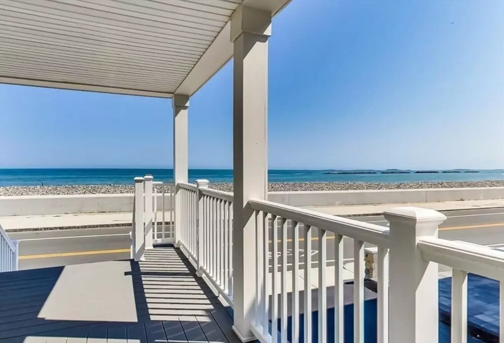 View from a porch with white railings overlooking the serene road, beach, and ocean on a clear day in Winthrop, MA.