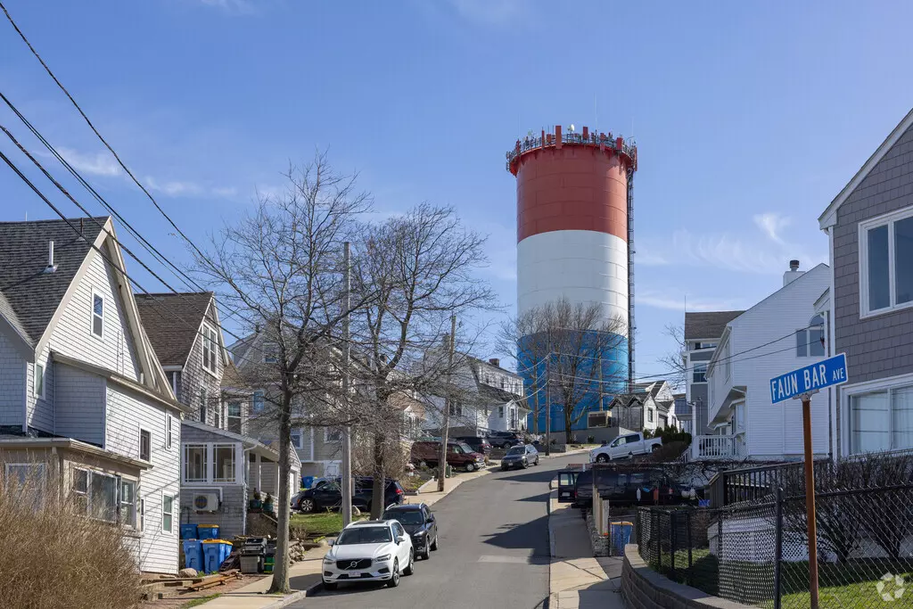 In Winthrop, MA, a residential street offers a glimpse of the tall, cylindrical water tower painted in red, white, and blue. Several houses line the tranquil street under a clear blue sky.