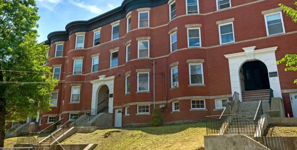 A row of four-story red brick apartment buildings in Winthrop, MA, features arched entrances and multiple windows, set gracefully on a grassy slope under a blue sky.