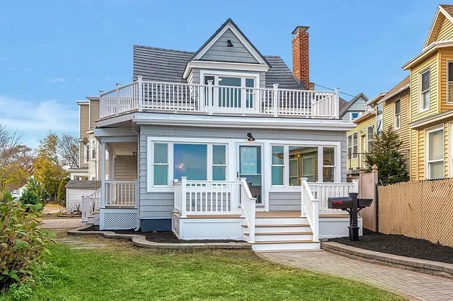 Nestled in the charming town of Winthrop, MA, this two-story gray house with white trim boasts a welcoming front porch and a second-story balcony. Set against a clear blue sky, it offers glimpses of neighboring homes, capturing the essence of New England living.