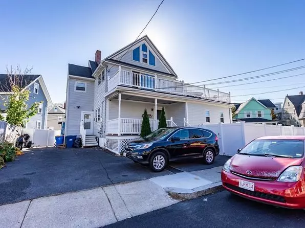 This charming two-story house in Winthrop, MA, features white siding, a welcoming porch, and a balcony. A black SUV and red sedan are parked neatly in the driveway. The surrounding blue sky and neighboring houses complete this idyllic neighborhood scene.