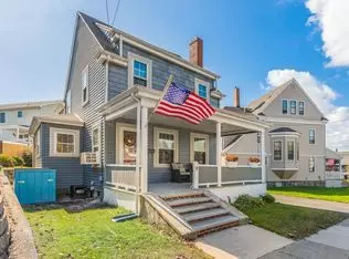 A charming two-story house in Winthrop, MA, features gray siding and a welcoming front porch displaying an American flag. Steps lead up to the entrance, set against a backdrop of similar homes under a picturesque blue sky with clouds.