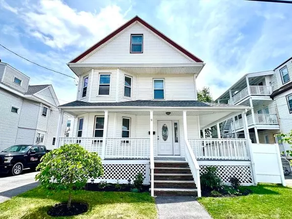 In picturesque Winthrop, MA, stands a charming white two-story house with a third attic level. It features a welcoming front porch and a cozy garden. On the left, the driveway cradles a parked car, completing this serene New England scene.