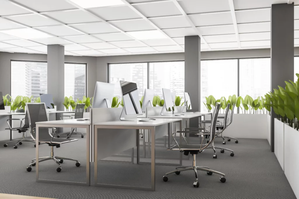 Modern office space with rows of desks and computers, gray swivel chairs, and planters with green plants. Large windows show a cityscape view. A reliable cleaning service ensures the environment stays pristine and welcoming.