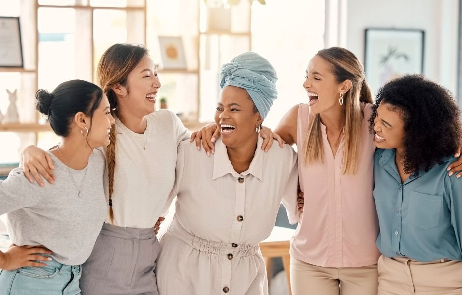 Five women laughing together, sharing cleaning tips for working women, and standing with arms around each other in a brightly lit room. Cleaning Tips for Working Women