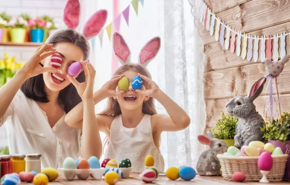 A woman and a girl wearing bunny ears hold decorated Easter eggs over their eyes at a table filled with Easter decorations, celebrating after finishing their Boston Marathon Week cleaning.