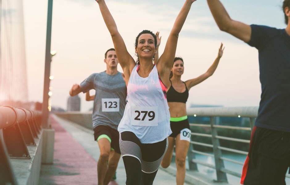 A woman wearing bib number 279 crosses the finish line with her arms raised, ahead of other runners, during a Boston Marathon Week Cleaning race on a bridge.