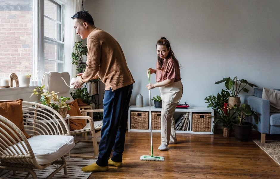 Two people tackle their Boston spring cleaning guide: one waters the lush indoor plants while the other diligently mops the wooden floor. Sunlight floods through large windows, illuminating cozy chairs and a freshened space ready for the season's renewal.