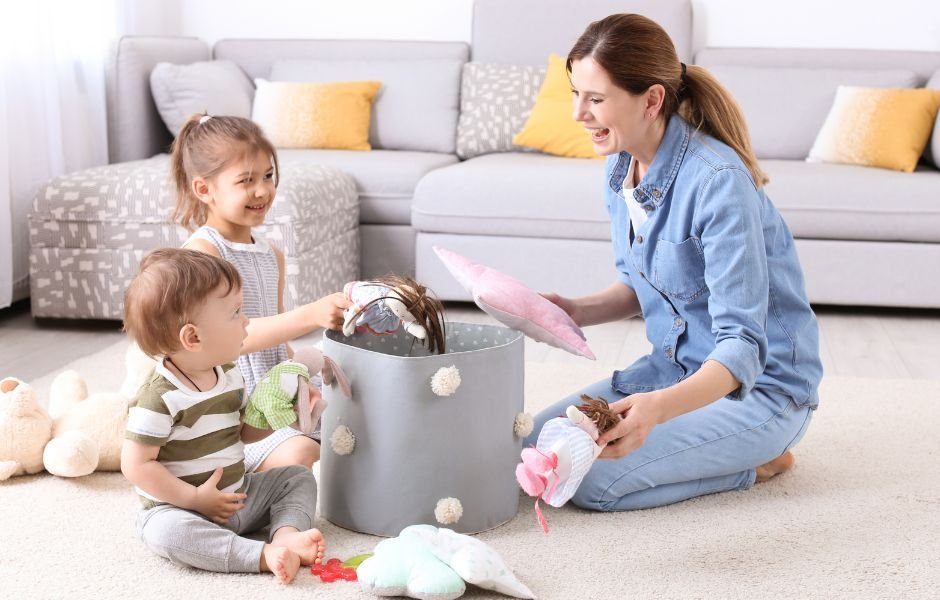 Woman and two young children sit on a carpeted floor, smiling as they engage in Spring Cleaning with Kids, putting stuffed toys into a large storage bin in the living room.