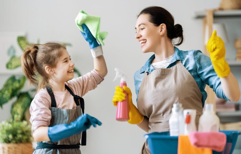A woman and a girl wearing aprons and rubber gloves smile and clean together with spray bottles and cloths in a bright room, enjoying an end-of-school-year cleaning for a fresh start summer. end-of-school-year cleaning